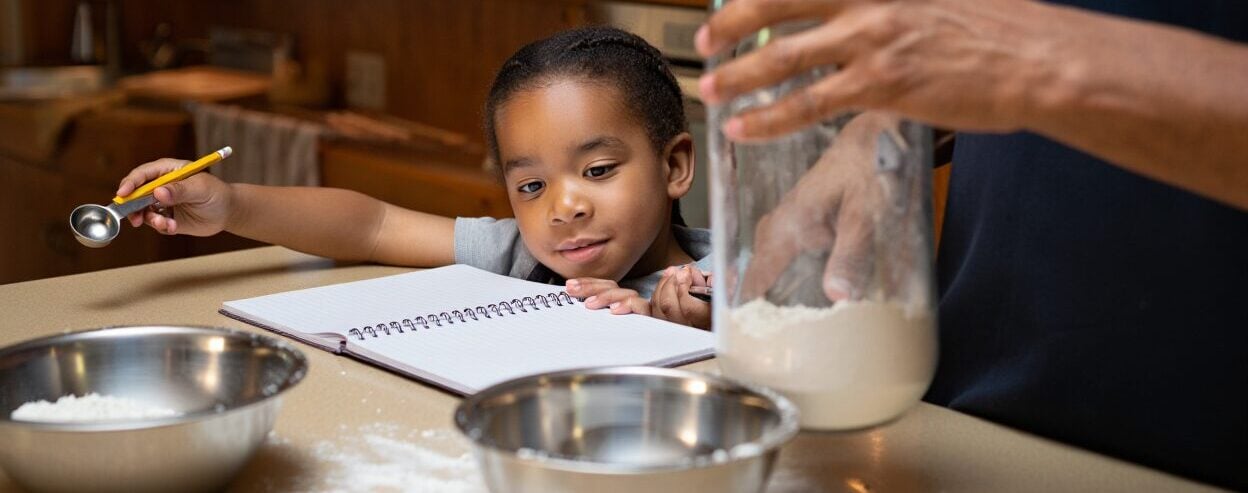 Child holding a measuring spoon and looking at a notebook while an adult bakes.