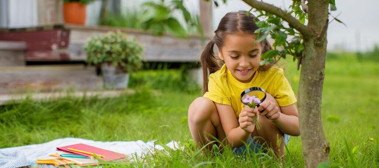 Child examining pink flowers with a magnifying glass in green grass.