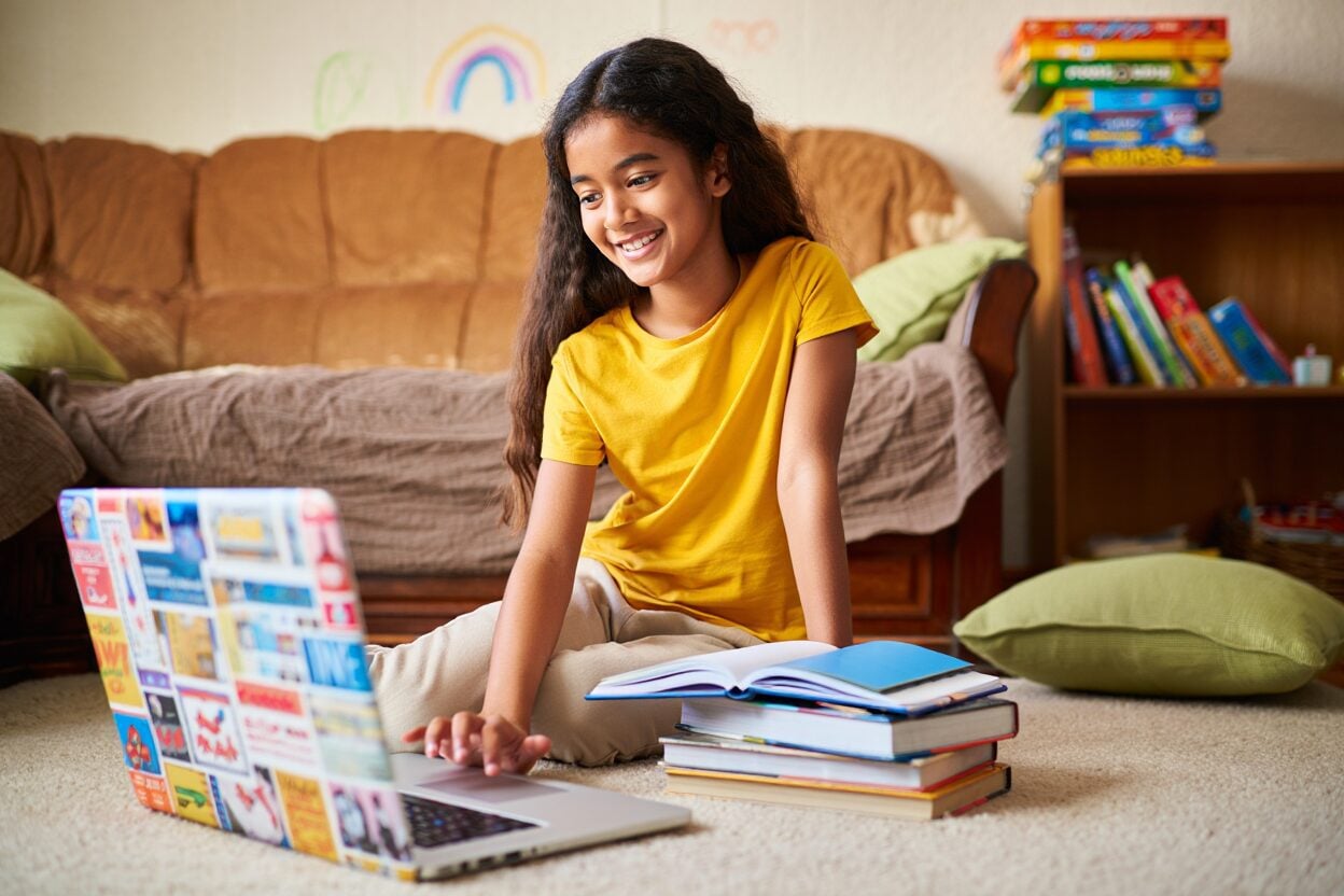 Smiling child on floor using laptop with books nearby.