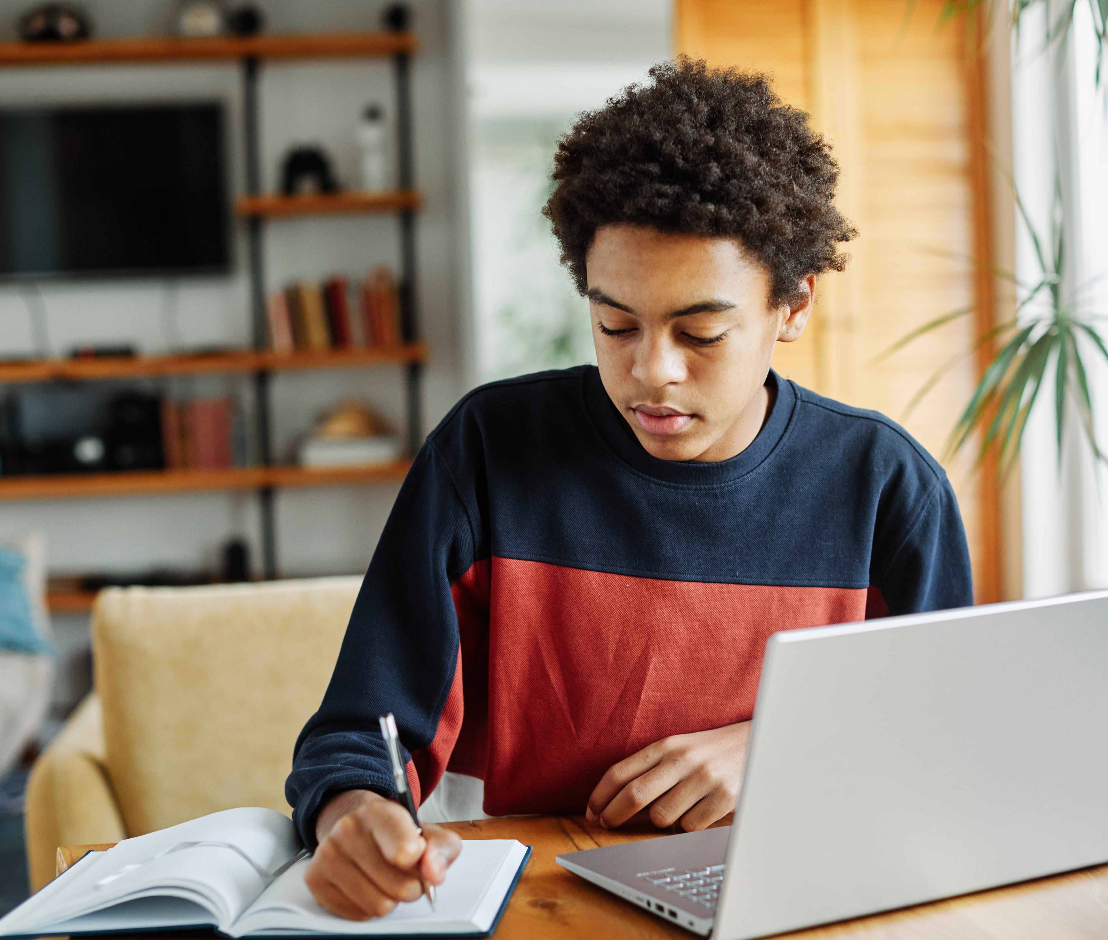 Young person writing in a notebook with a laptop nearby.